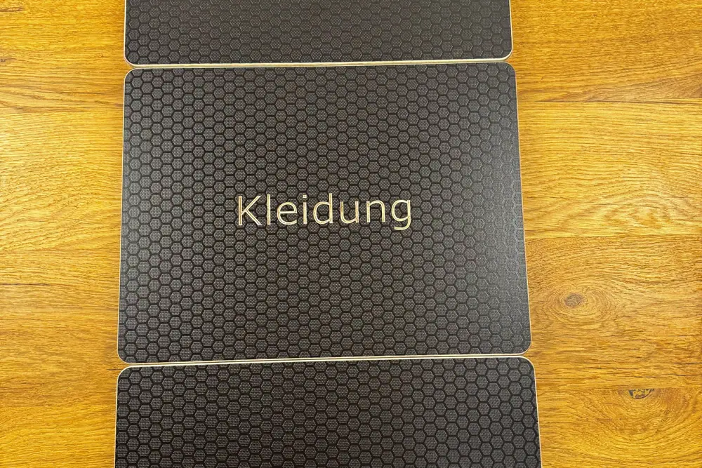Drei Eurobox-Deckel mit Hexagon-Oberfläche, in die die Wörter Werkzeug, Kleidung und Essen gefräst sind, auf einem Holztisch